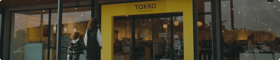 A shopfront with a bright yellow sign that reads TAKKO FASHION above the entrance, displaying clothing and mannequins inside a modern glass-fronted building.