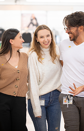 Three people stand close together indoors, smiling and laughing. They each wear name badges and appear to be colleagues enjoying a friendly moment. The background is blurred, suggesting a casual workplace environment.