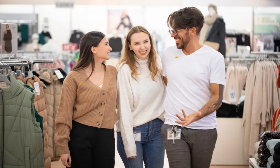 Three people, two women and one man, smiling and talking together in a clothes shop, surrounded by rails of clothes. They appear to be colleagues, each wearing name badges.