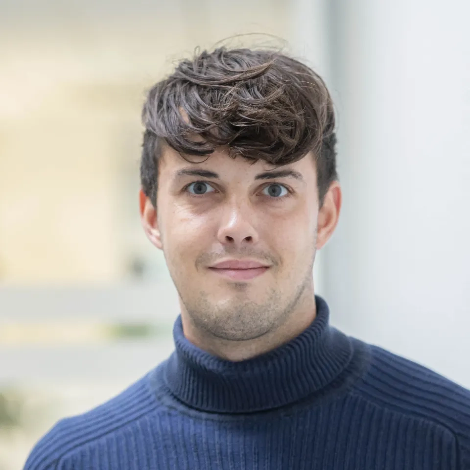 A young man with short, wavy brown hair and light skin wears a dark blue polo-neck jumper and looks at the camera with a neutral expression against a blurred indoor background.