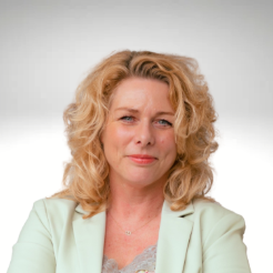 A woman with curly blonde hair wearing a light-coloured blazer smiles softly whilst looking at the camera, set against a plain, gradient background.