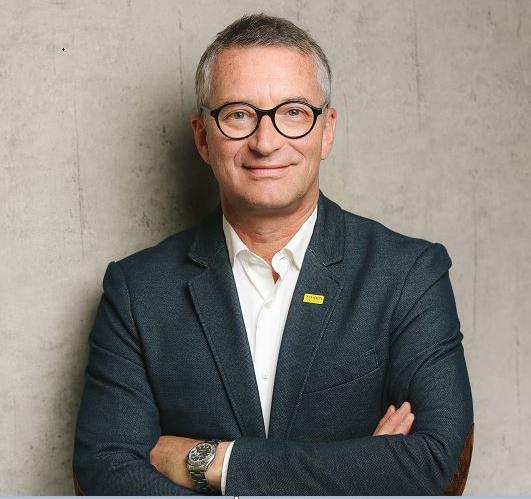 A middle-aged man with short grey hair and glasses, wearing a dark blazer over a white shirt, stands with arms folded and smiles in front of a textured beige wall.