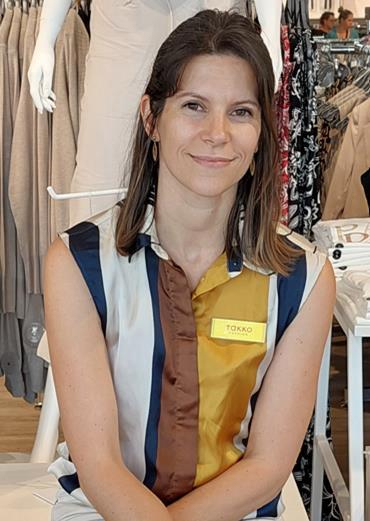 A woman with straight brown hair, wearing a sleeveless striped blouse and a yellow name badge reading Takko, sits and smiles inside a clothes shop with racks of clothes visible in the background.