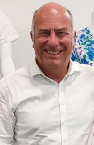 A middle-aged man with a bald head is smiling and wearing a white button-up shirt. There are mannequins dressed in light-coloured clothing and a floral print top in the background.