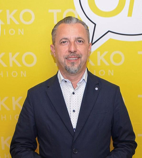 A man with short grey hair and a trimmed goatee, wearing a dark suit jacket and a light polka-dot shirt, stands in front of a yellow background with TOKKO FASHION and a speech bubble graphic.