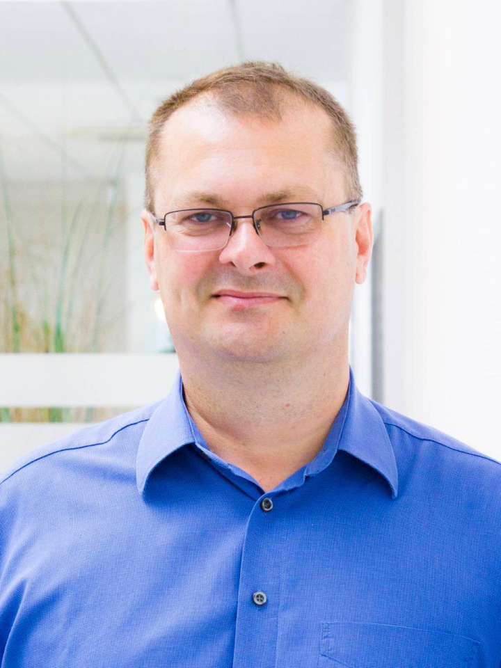 Ein Mann mit kurzen Haaren und Brille, der ein blaues Hemd mit Kragen trägt, steht in einem hell erleuchteten Raum mit weißen Wänden und Glasscheiben im Hintergrund.