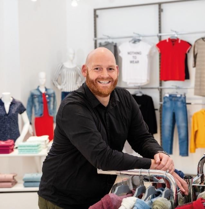 A smiling man with a beard stands in a clothes shop, leaning on rails with neatly folded clothes. Mannequins wearing casual outfits and shelves with colourful shirts and jeans are visible in the background.