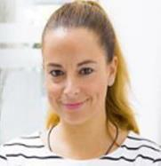 A woman with fair skin and long blonde hair tied back, smiling at the camera. She is wearing a white top with black stripes and is in a bright, indoor setting.