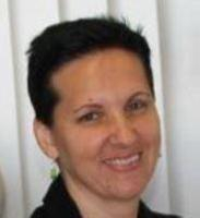 A person with short dark hair and light skin smiling, wearing a black top, and standing in front of a background with vertical white blinds.
