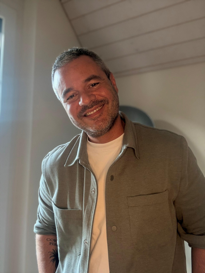 A man with short grey hair and a beard smiles warmly, wearing a light olive-green button-up shirt over a white T-shirt. He stands in a softly lit room with a slanted ceiling and a window to his left.