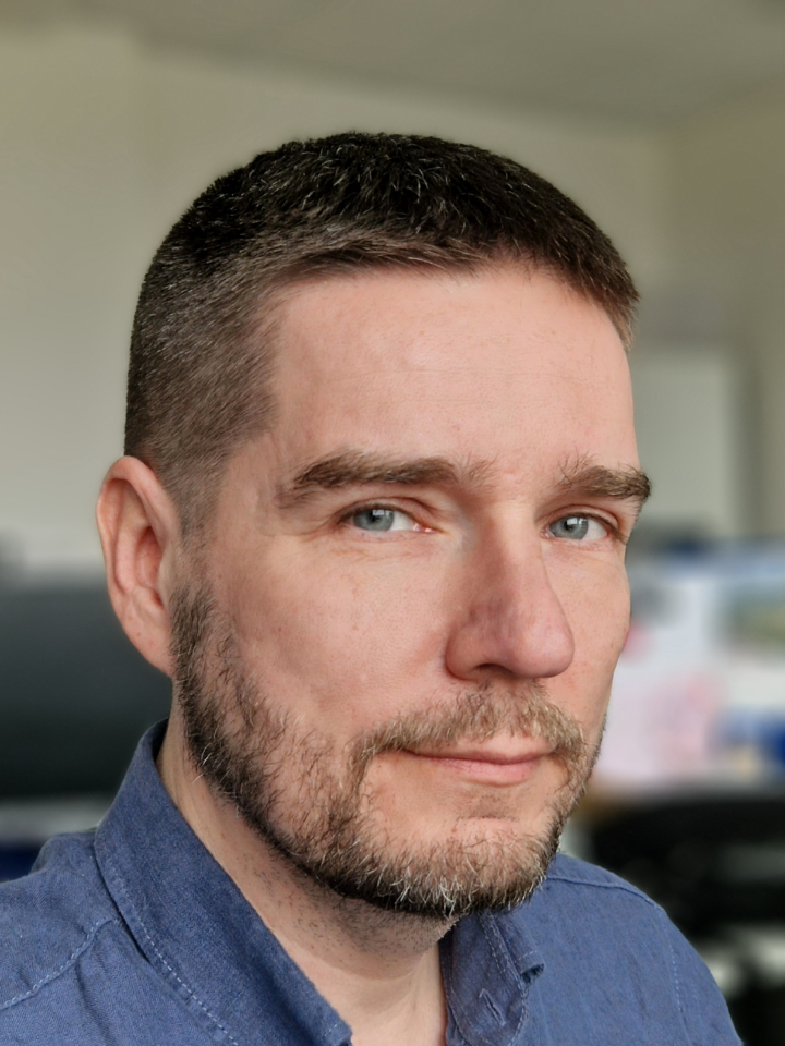 A man with short brown hair and a trimmed beard, wearing a blue collared shirt, looks at the camera in an indoor setting with a blurred office-like background.