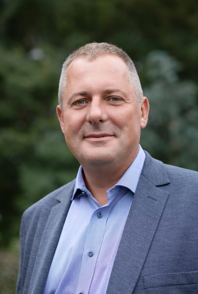 Ein Mann mittleren Alters mit kurzen Haaren, der ein blaues Hemd und einen strukturierten blauen Blazer trägt, steht im Freien vor einem unscharfen Hintergrund aus Grünpflanzen und lächelt leicht in die Kamera.