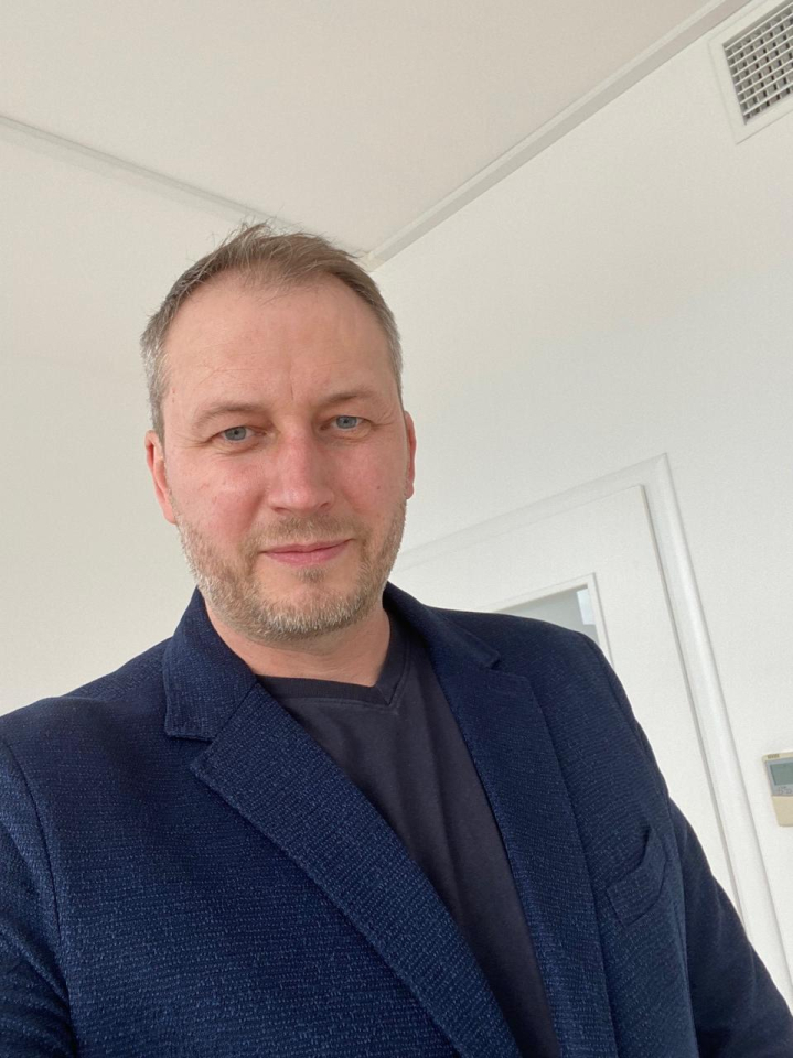 A man with short hair and a trimmed beard wears a navy blazer over a dark shirt, standing indoors with a white wall and door in the background.