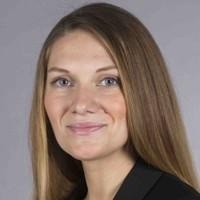 Eine Frau mit langem, glattem hellbraunem Haar und schwarzem Blazer lächelt sanft in die Kamera vor einem schlichten grauen Hintergrund.