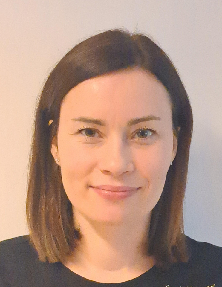 A woman with straight brown hair and fair skin smiles slightly at the camera, standing against a plain light background. She is wearing a dark top.
