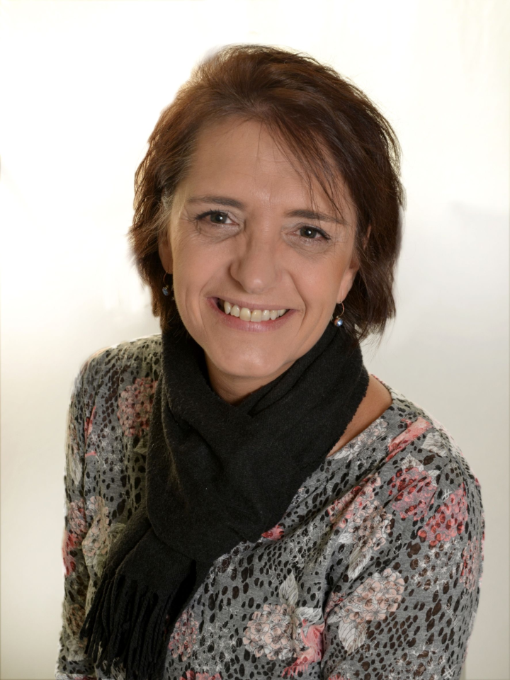 A woman with short brown hair smiles at the camera. She is wearing a floral-patterned top and a black scarf, with a light-coloured background behind her.