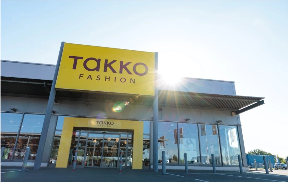 A Takko Fashion shop with a large yellow sign above the entrance on a sunny day. Sunlight is shining over the modern glass building, and the car park is visible in front.