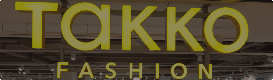 Yellow illuminated TAKKO FASHION sign inside a shop, with ceiling lights and structural elements visible in the background.